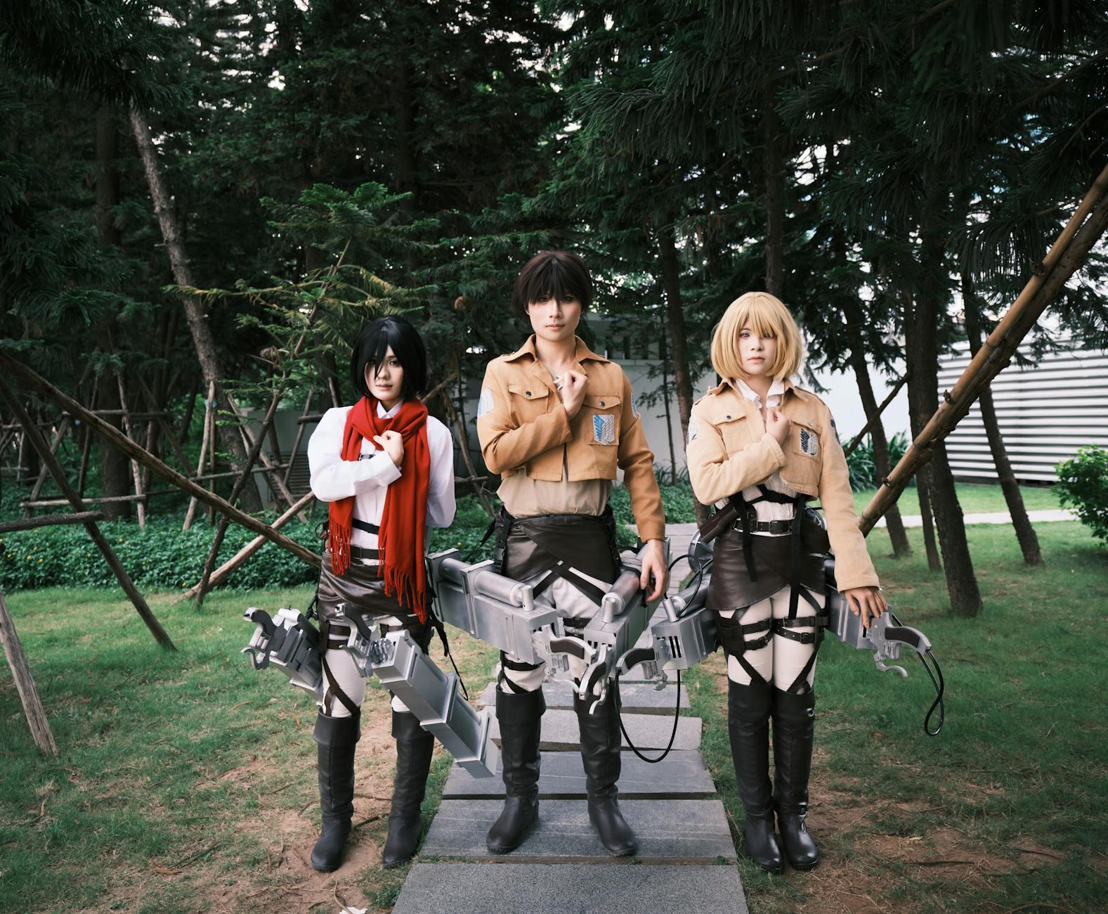 Three cosplayers in detailed anime costumes pose outdoors amidst trees.