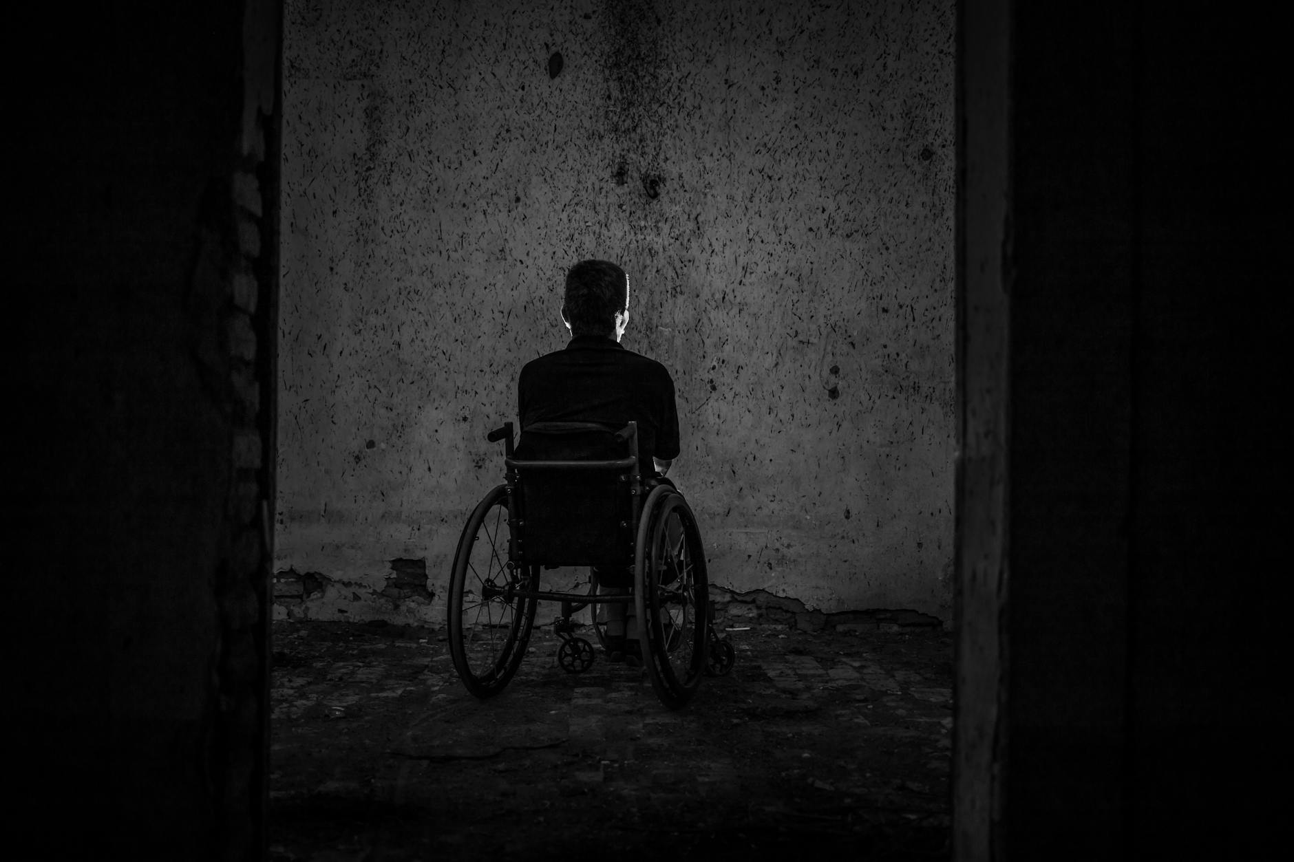 A man sits alone in a wheelchair facing a wall in a dark, empty room, evoking solitude.