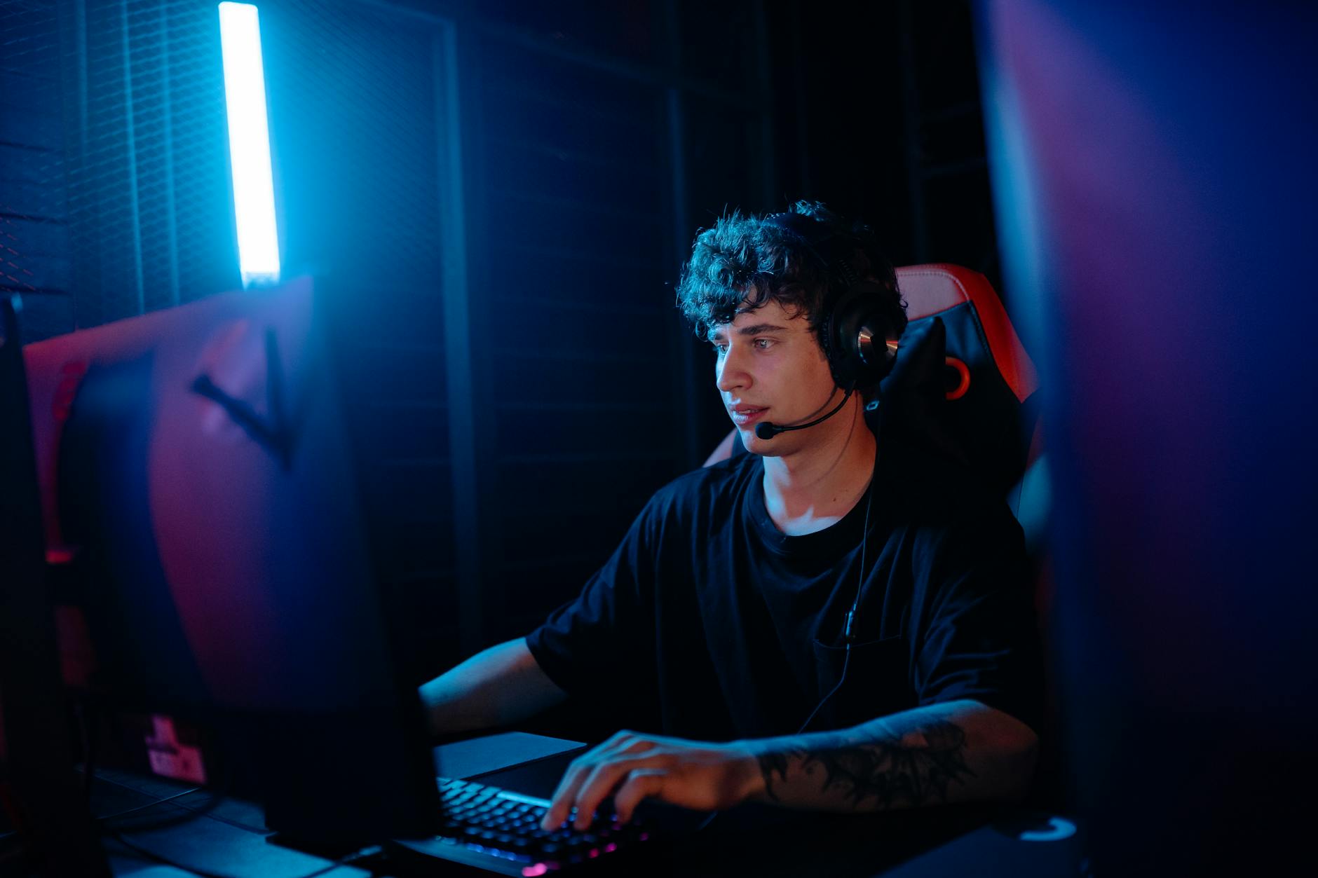A young man intensely gaming with a headset at a computer desk, illuminated by blue light.