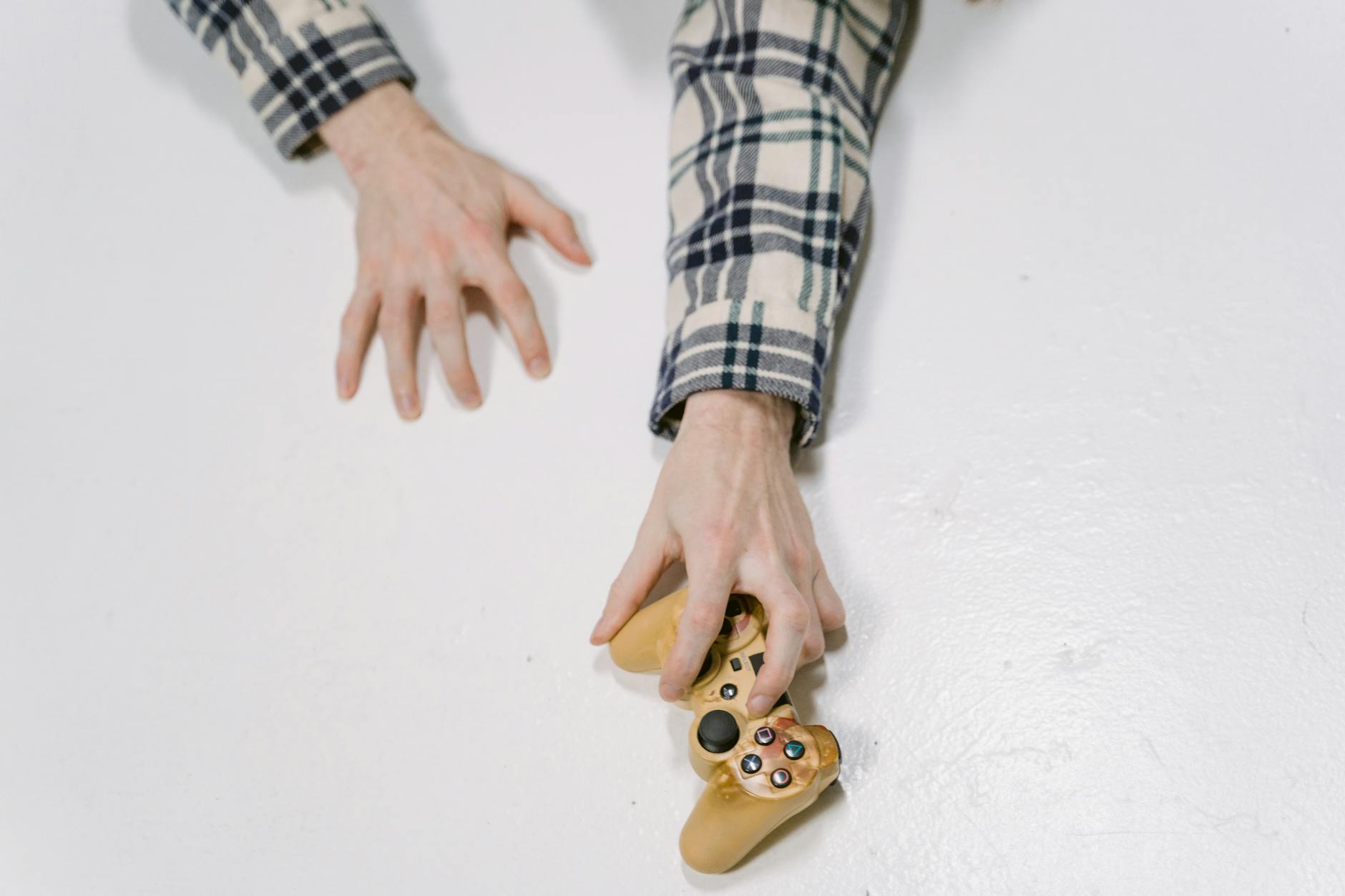 A person reaching for a video game controller on a white surface, wearing a plaid shirt.