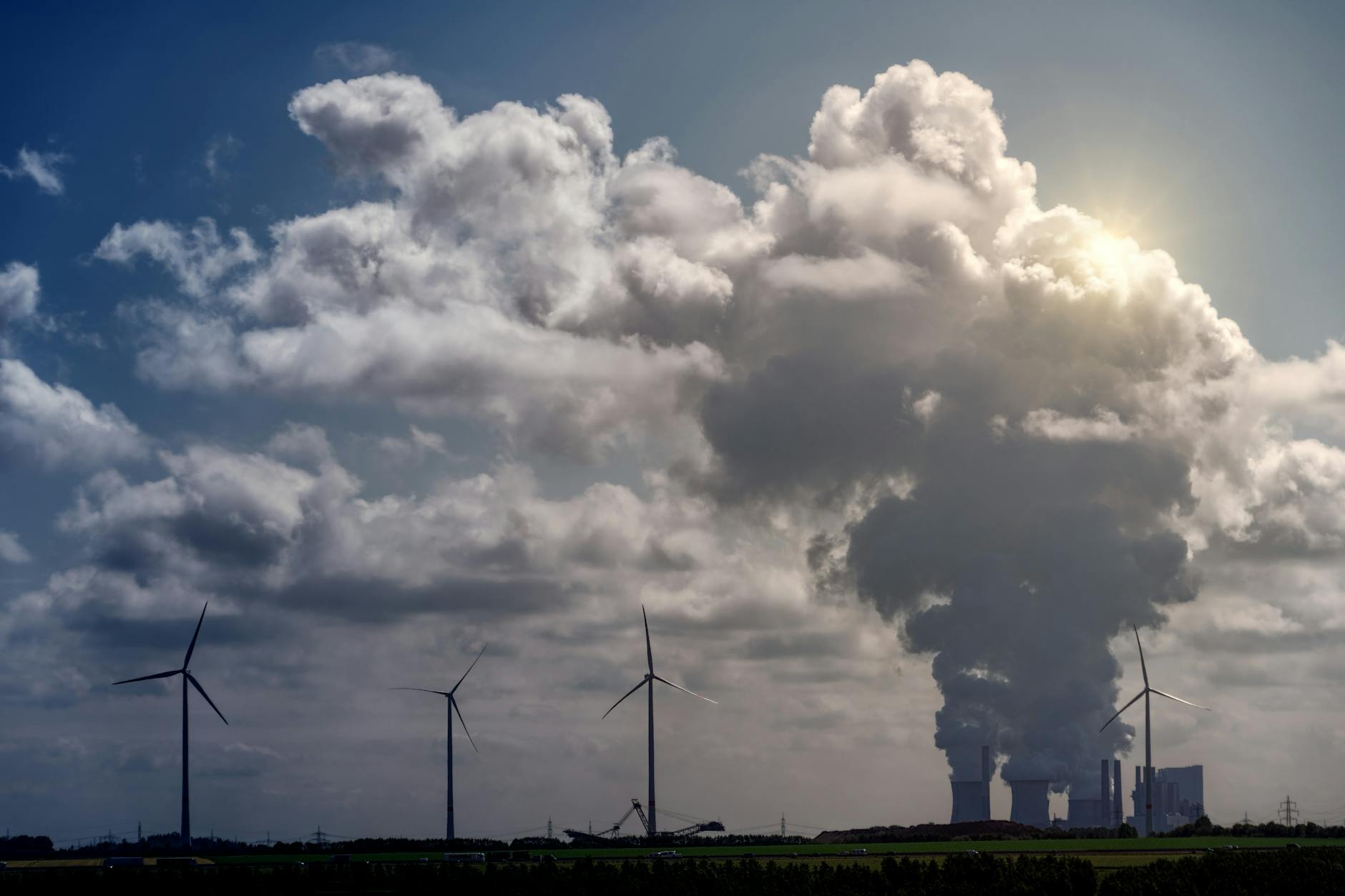 Wind turbines and a power plant under dramatic cumulus clouds in a rural landscape.