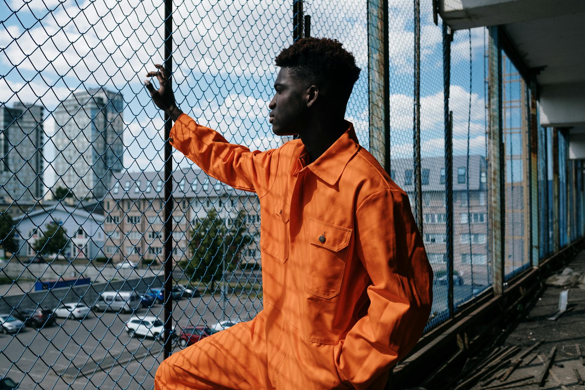 Man in orange overalls leans against a chain-link fence, cityscape in background.