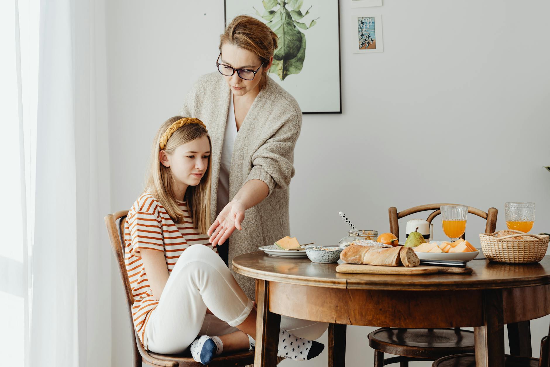 A mother guides her teenage daughter in dining etiquette at a cozy indoor setting.