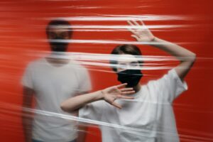 Two adults wearing masks stand behind a plastic sheet, symbolizing isolation and protection.