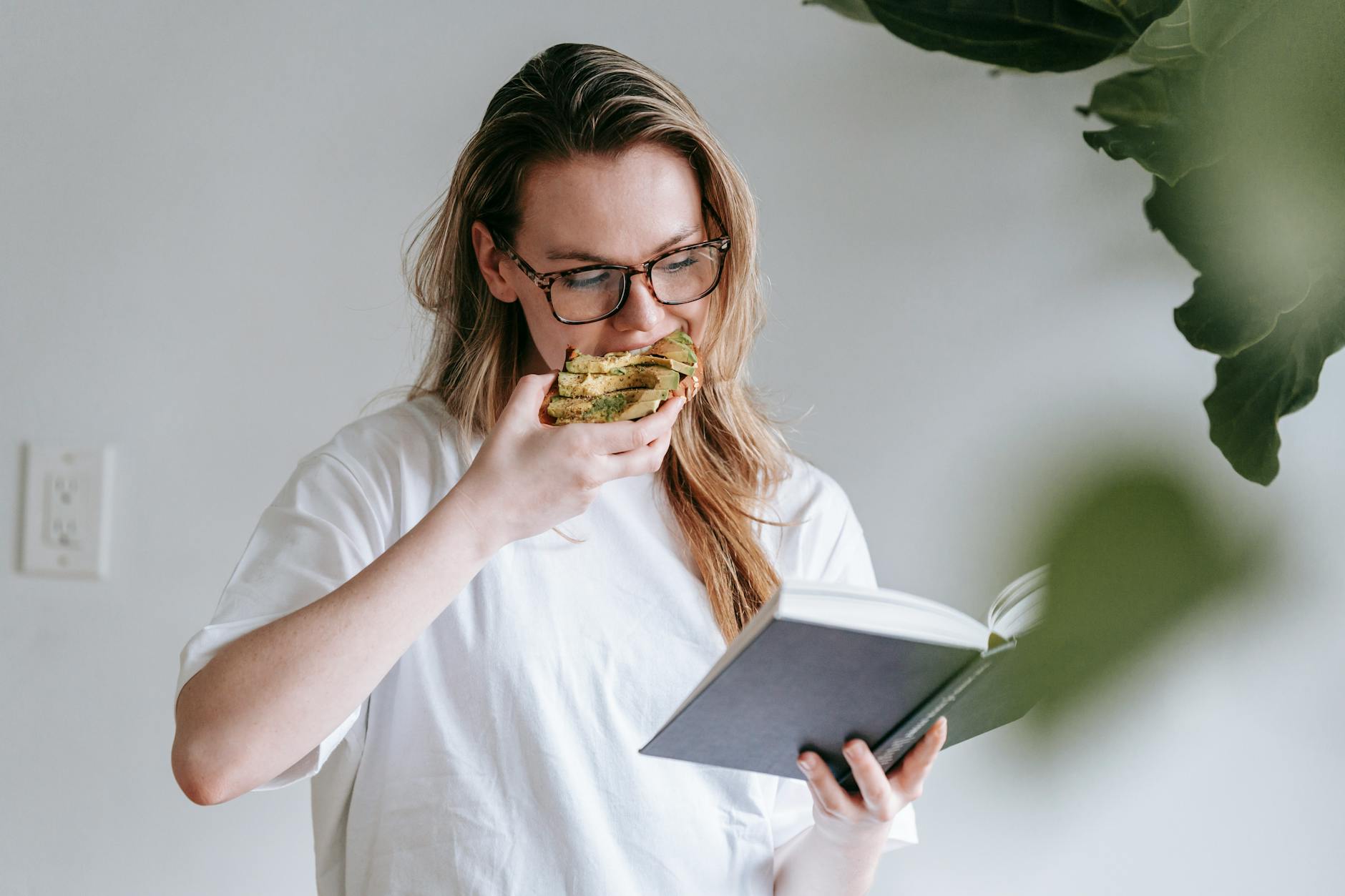A young woman reading a book and eating a sandwich, enjoying leisure time indoors.