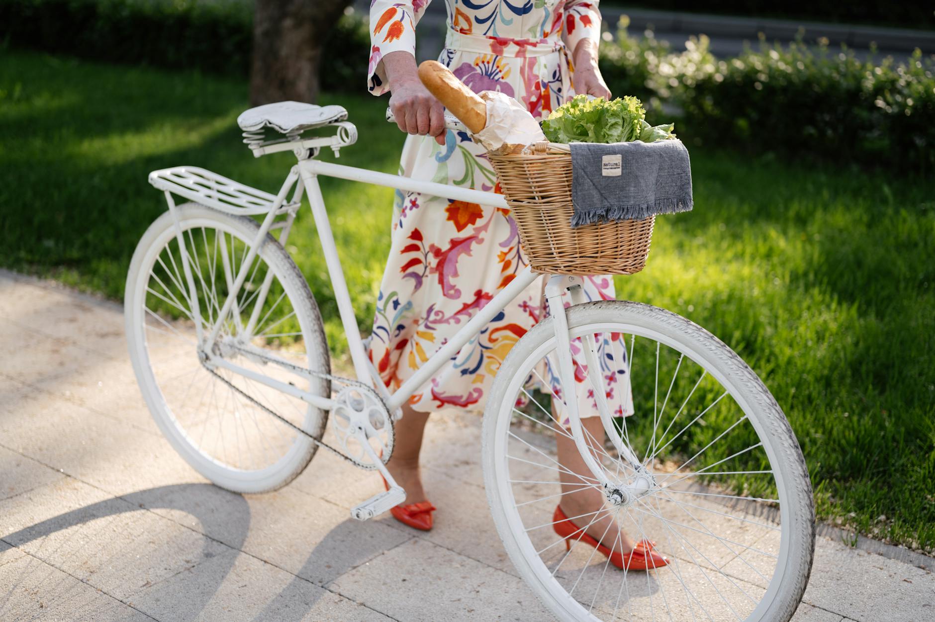 Woman in vibrant dress walking bicycle with basket full of bread and greens, showcasing trendy fashion outdoors.