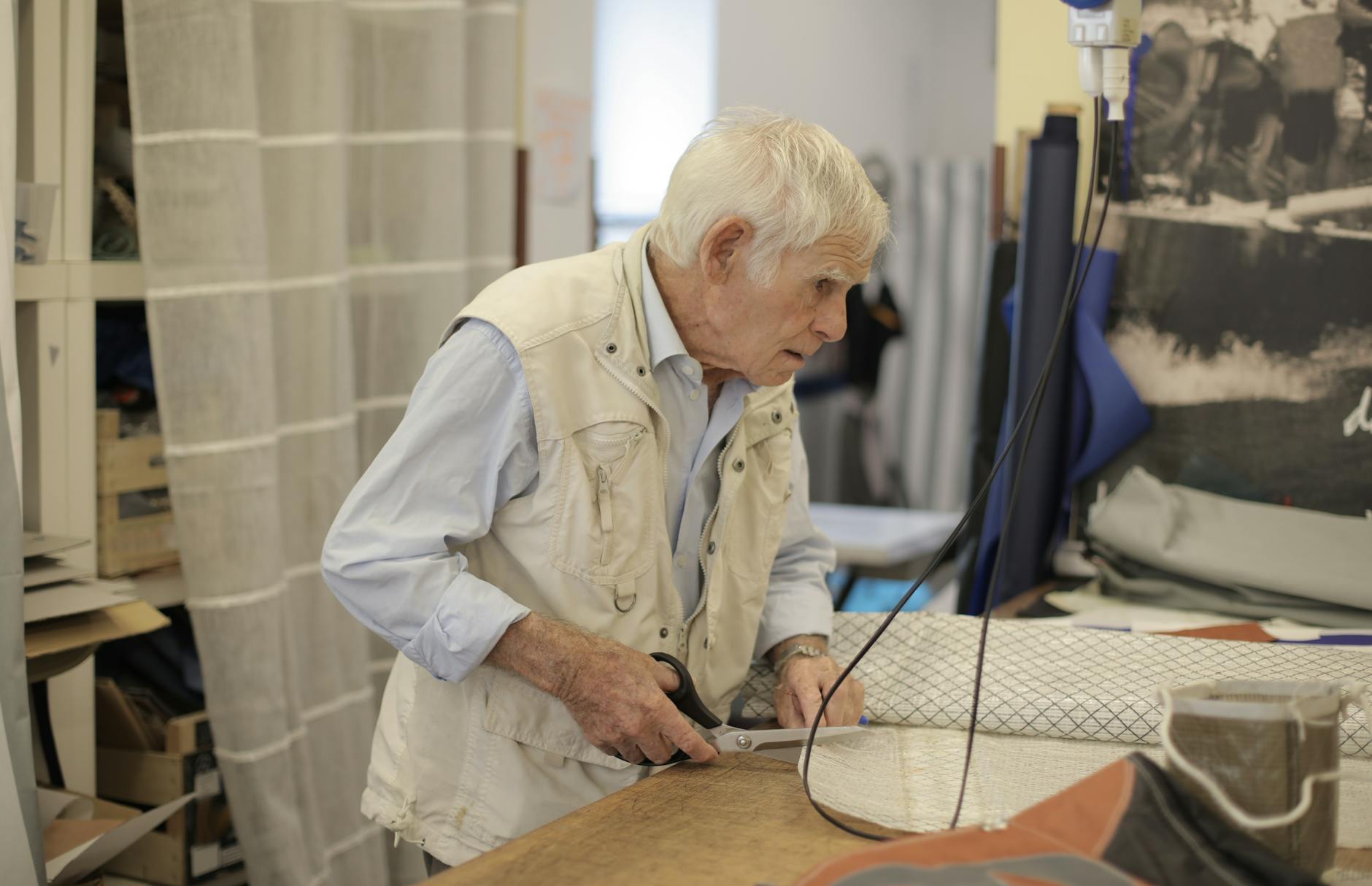 An elderly tailor with white hair cuts fabric at a workshop table, showcasing skill and concentration.