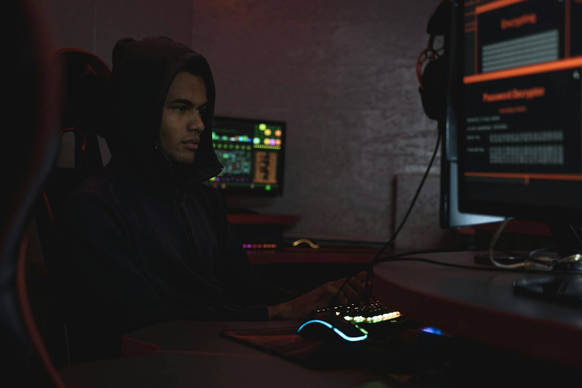 A hacker in a hoodie working in a dimly lit room, focusing on cyber security tasks on multiple monitors.