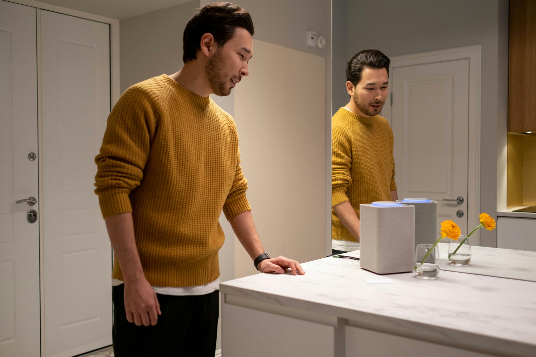 A man in a sweater reflects in a mirror next to a smart speaker in a modern home setting.
